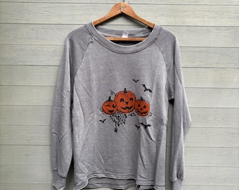 May include: A grey long-sleeve sweatshirt featuring a graphic print of three orange jack-o'-lanterns, a black spiderweb, and several black bats. The garment has a crew neckline, raglan sleeves, and a raw, unfinished hemline at the bottom.