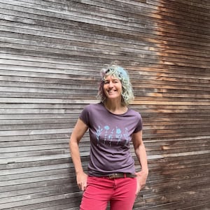 Op de afbeelding: Een persoon draagt een paars t-shirt met een bloemenontwerp en een rode broek. Het t-shirt heeft een ronde hals en korte mouwen. De broek is effen van kleur en heeft een riem. De achtergrond is een houten muur.