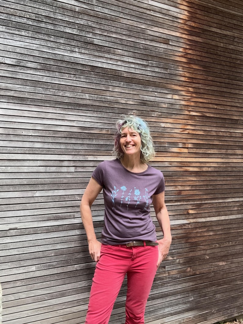 Op de afbeelding: Een persoon draagt een paars t-shirt met een bloemenontwerp en een rode broek. Het t-shirt heeft een ronde hals en korte mouwen. De broek is effen van kleur en heeft een riem. De achtergrond is een houten muur.