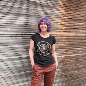 May include: A woman with purple hair is wearing a black t-shirt with a gold mandala design. She is standing in front of a wooden wall.