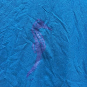 Pu&ograve; includere: Una maglietta blu turchese con un disegno di cavalluccio marino viola. Il cavalluccio marino &egrave; stampato sulla parte anteriore della maglietta. Il tessuto &egrave; leggermente stropicciato, dando alla maglietta un aspetto casual.