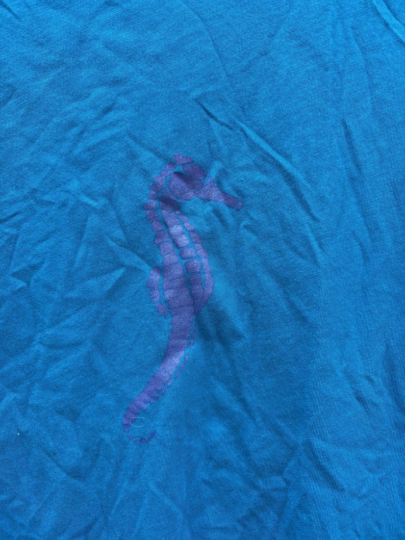 Pu&ograve; includere: Una maglietta blu turchese con un disegno di cavalluccio marino viola. Il cavalluccio marino &egrave; stampato sulla parte anteriore della maglietta. Il tessuto &egrave; leggermente stropicciato, dando alla maglietta un aspetto casual.