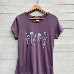Op de afbeelding: Een pruimkleurig T-shirt met korte mouwen, versierd met een lichtblauw bloemmotief. Het ontwerp bevat verschillende gestileerde bloemen en stelen. Het shirt hangt aan een houten hanger tegen een lichtgekleurde houten achtergrond.