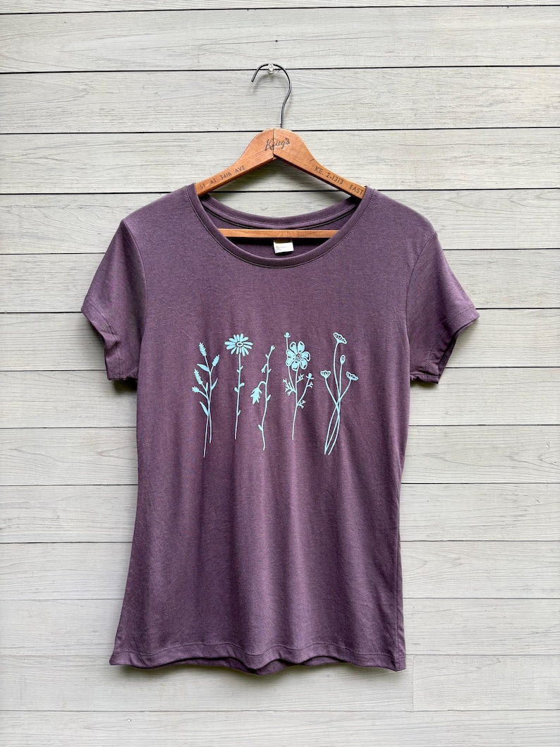 Op de afbeelding: Een pruimkleurig T-shirt met korte mouwen, versierd met een lichtblauw bloemmotief. Het ontwerp bevat verschillende gestileerde bloemen en stelen. Het shirt hangt aan een houten hanger tegen een lichtgekleurde houten achtergrond.