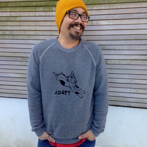 May include: A gray sweatshirt with a black graphic of a dog and the word "ADOPT" printed below. The person is wearing a yellow beanie, glasses, and blue jeans.