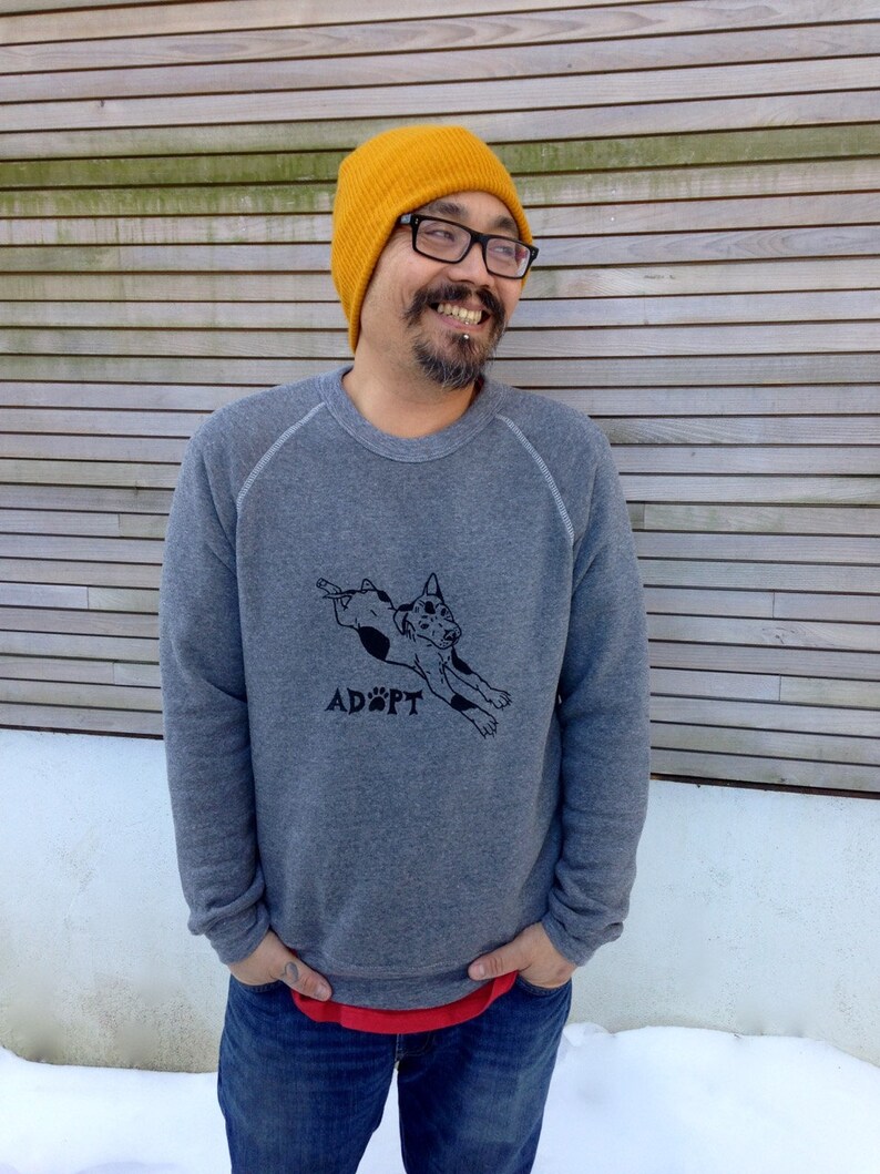 May include: A gray sweatshirt with a black graphic of a dog and the word "ADOPT" printed below. The person is wearing a yellow beanie, glasses, and blue jeans.