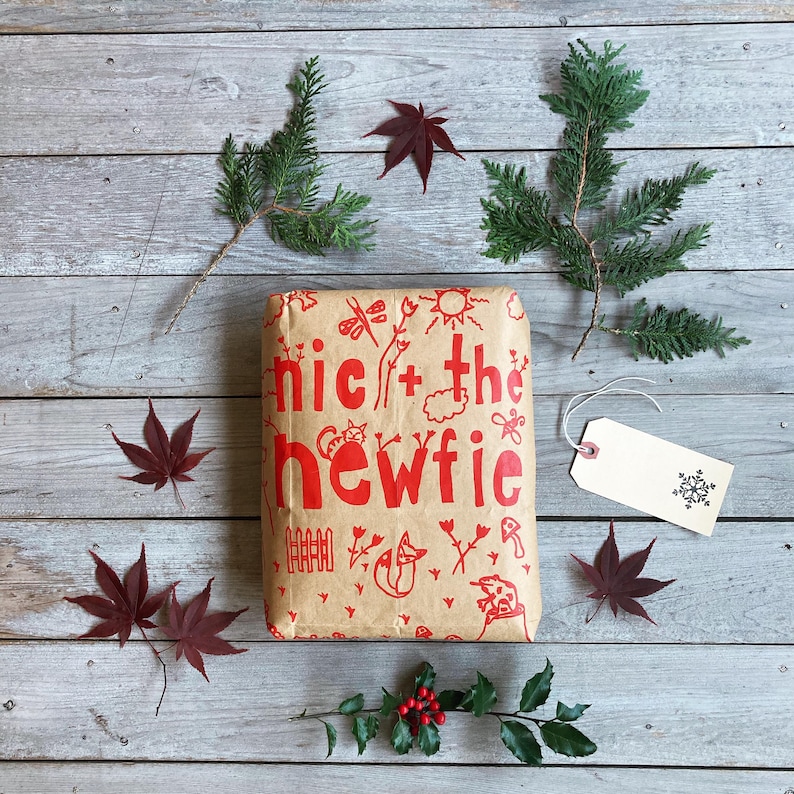 Op de afbeelding: Een cadeau verpakt in bruin papier met rode illustraties en tekst, waaronder "nic + the newfie". Het is omgeven door rode bladeren, groene bladeren en een label met een sneeuwvlokontwerp.