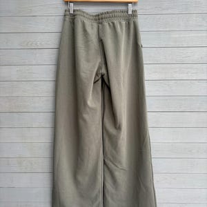 K&ouml;nnte beinhalten: Olivgr&uuml;ne Hose mit weitem Bein und elastischem Bund mit gerafftem Abschluss. Die Hose ist aus einem einfarbigen Stoff gefertigt und h&auml;ngt an einem Holzkleiderb&uuml;gel vor einer Wand mit hellgrauer Holzvert&auml;felung.