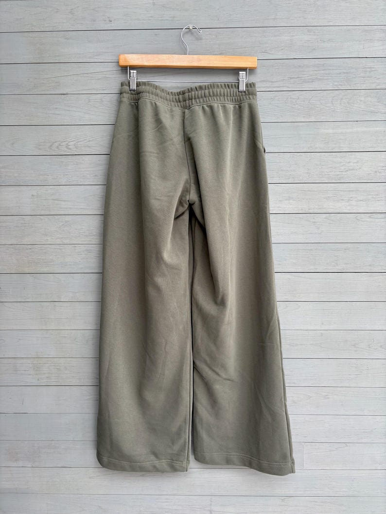 K&ouml;nnte beinhalten: Olivgr&uuml;ne Hose mit weitem Bein und elastischem Bund mit gerafftem Abschluss. Die Hose ist aus einem einfarbigen Stoff gefertigt und h&auml;ngt an einem Holzkleiderb&uuml;gel vor einer Wand mit hellgrauer Holzvert&auml;felung.