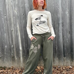 K&ouml;nnte beinhalten: Ein cremefarbenes Langarmshirt mit einer schwarzen Grafik von zwei V&ouml;geln und eine olivgr&uuml;ne Jogginghose mit wei&szlig;en Farn-Aufdrucken am Bein, ausgestattet mit einem elastischen Bund mit Kordelzug und Seitentaschen.
