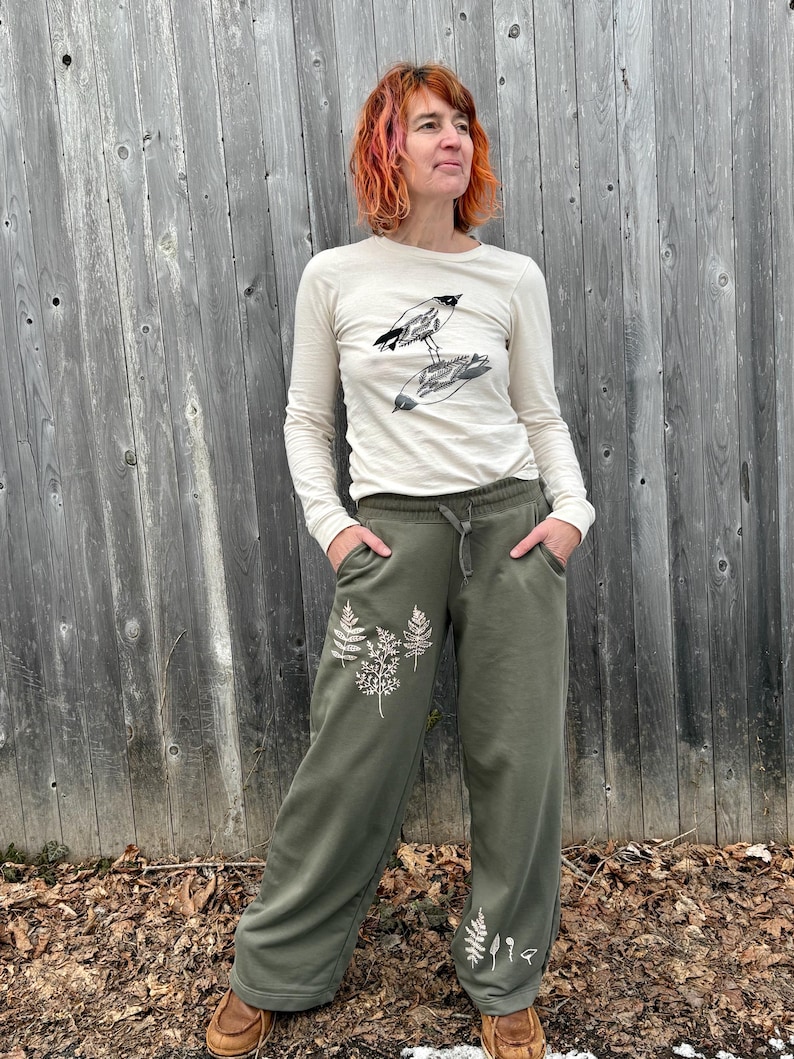 K&ouml;nnte beinhalten: Ein cremefarbenes Langarmshirt mit einer schwarzen Grafik von zwei V&ouml;geln und eine olivgr&uuml;ne Jogginghose mit wei&szlig;en Farn-Aufdrucken am Bein, ausgestattet mit einem elastischen Bund mit Kordelzug und Seitentaschen.