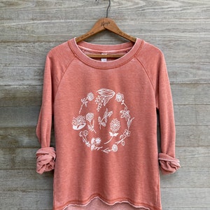 May include: A coral colored sweatshirt with a white floral design in a circle on the front. The sweatshirt has long sleeves and a relaxed fit.