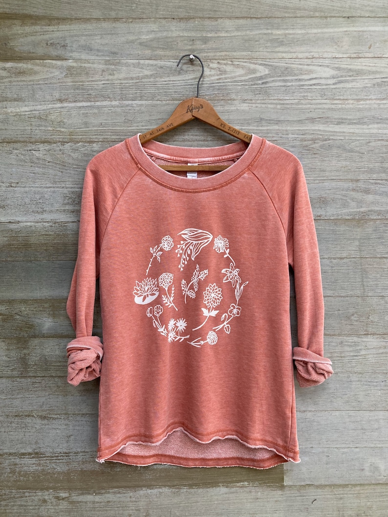 May include: A coral colored sweatshirt with a white floral design in a circle on the front. The sweatshirt has long sleeves and a relaxed fit.
