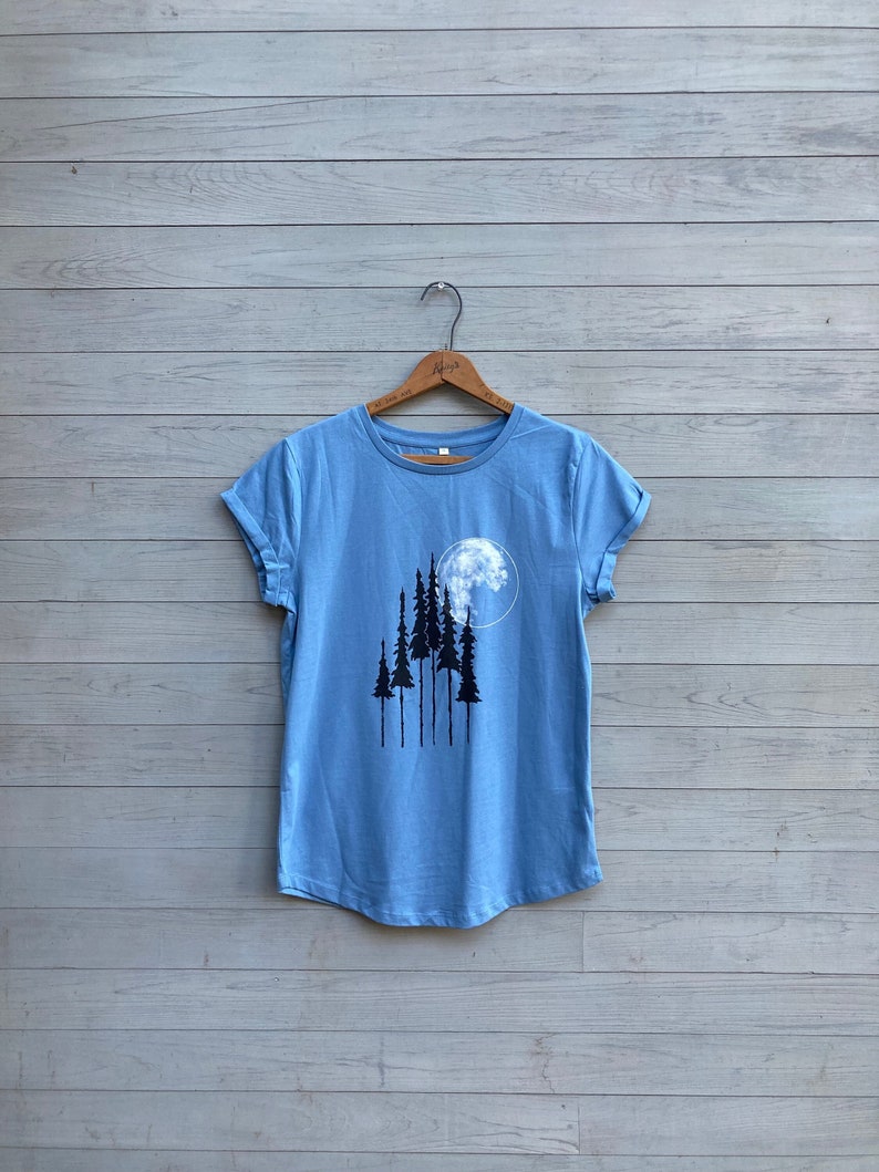 Puede incluir: Una camiseta azul claro con un gr&aacute;fico de una luna y &aacute;rboles en negro. La camiseta tiene las mangas enrolladas y un dobladillo curvo.