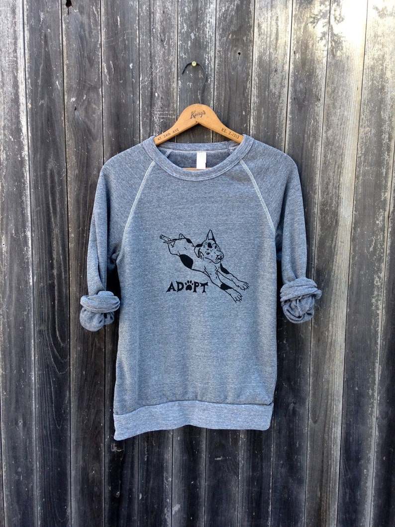 May include: A heather gray sweatshirt with a black graphic of a dog leaping and the word "ADOPT" printed below. The long sleeves are rolled up. The sweatshirt is hanging on a wooden hanger against a weathered wood background.