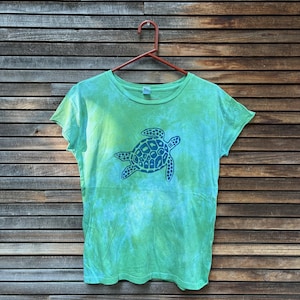 May include: A green tie-dye t-shirt with a blue sea turtle graphic. The shirt is hanging on a red hanger against a wooden plank background. The t-shirt has a crew neck and short sleeves.