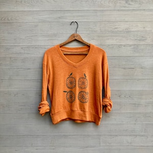 May include: Orange V-neck sweatshirt with a black graphic of four orange slices. The long-sleeved shirt is hanging on a wooden hanger against a light grey wooden background. The sleeves are rolled up.