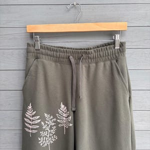 K&ouml;nnte beinhalten: Olivgr&uuml;ne Jogginghose mit elastischem Bund und Kordelzug sowie einer wei&szlig;en Linien-Grafik von drei verschiedenen Farnbl&auml;ttern auf dem linken Hosenbein. Die Jogginghose h&auml;ngt an einem Holzkleiderb&uuml;gel mit Metallklammern.