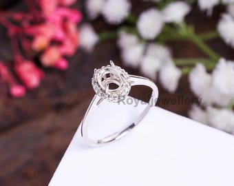 May include: A silver ring with a halo setting, featuring a central space for a gemstone surrounded by small, sparkling stones. The ring's band is slender and polished, with the words "Royal Jewellery" visible.