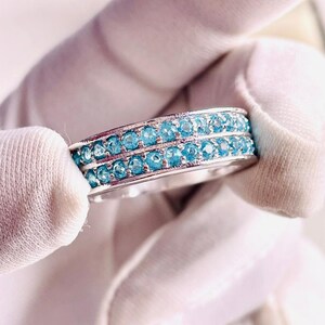 Può includere: Un anello d'argento con due file di piccole gemme rotonde blu chiaro. L'anello è tenuto tra due mani guantate di bianco, mostrando il design e il colore. Le gemme sono uniformemente distanziate.