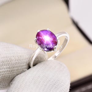 May include: A silver ring with a large, oval star ruby gemstone. The ruby is a deep purple colour with a star effect. The ring's band is a simple, polished silver design.