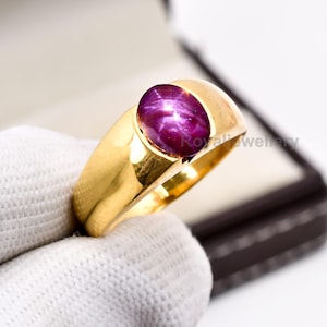 Może przedstawiać: Złoty pierścionek z dużym, owalnym, głęboko czerwonym kamieniem szlachetnym z efektem gwiazdy. Pierścionek ma grubą, polerowaną obrączkę i klasyczny design. Kamień szlachetny jest centralnym punktem, prezentując swój bogaty kolor.