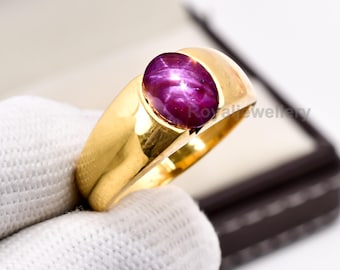May include: A gold ring with a large, oval, deep red gemstone displaying a star effect. The ring has a thick, polished band and a classic design. The gemstone is the focal point, set in the centre, showcasing its rich colour and unique pattern.