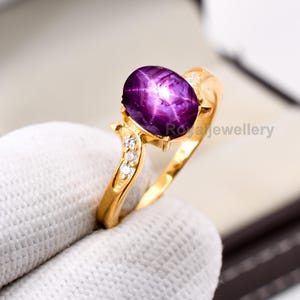 May include: A gold ring with a large, oval, deep purple gemstone displaying a star pattern. The ring is embellished with small, clear gemstones. The text "Royal Jewellery" is visible on the ring.