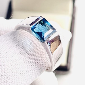 Peut inclure: Une bague en argent ornée d'une pierre précieuse bleue carrée et vibrante. La bague présente un design poli et moderne, mettant en valeur la pierre. La bague est présentée sur un fond blanc, soulignant ses détails.