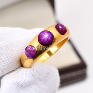 May include: A gold ring featuring three round, deep purple gemstones. The ring has a smooth, polished band and the gemstones are evenly spaced across the top. The ring is displayed in a jewelry box.