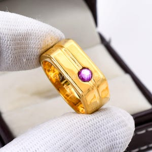 Puede incluir: Un anillo dorado con un diseño rectangular y una pequeña gema redonda de color púrpura. El anillo está sostenido por una mano enguantada, y el fondo es una caja oscura. El anillo tiene un acabado pulido.
