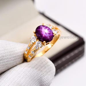 May include: A gold ring featuring a large, oval, deep purple gemstone with a star-like effect. The ring is accented with smaller, clear gemstones and has a split shank design. The ring is presented in a jewelry box.