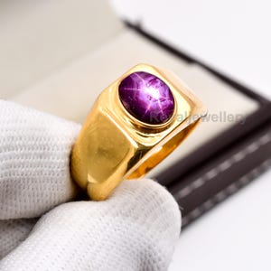Puede incluir: Un anillo de oro con una gran gema de rubí estrella ovalada. El rubí es de color púrpura oscuro con un efecto de estrella visible. El anillo tiene una banda rectangular clásica y un acabado pulido.