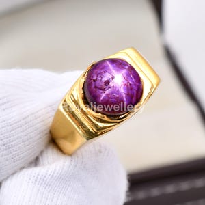 May include: A gold ring featuring a large, round, purple gemstone with a star-like effect. The ring has a thick, rectangular band and a bezel setting. The gemstone is a deep, rich purple color. The ring is displayed on a white surface.