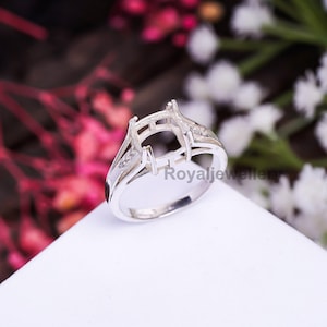 May include: A silver ring setting with a detailed design, featuring a space for a central stone and smaller accent stones. The ring is displayed on a white surface with a blurred floral background, showcasing the intricate metalwork and overall elegance.