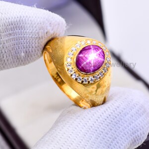 May include: A gold ring featuring a large, oval, purplish-pink gemstone with a star-like effect. The gemstone is surrounded by a halo of small, clear gemstones. The ring is held by a person wearing a white glove.
