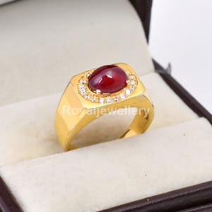 May include: A gold ring featuring a large, oval, deep red gemstone, encircled by smaller, clear stones. The ring has a rectangular face and a polished band. The text "Royaljewellery" is visible on the ring. A luxurious piece of jewellery.