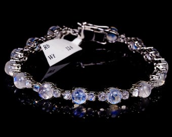 Puede incluir: Una pulsera de plata con múltiples piedras lunares redondas e iridiscentes. La pulsera está sobre un fondo negro, resaltando los tonos azules y blancos de las piedras. Una etiqueta con el texto "Rb" y "Wt" está adjunta.