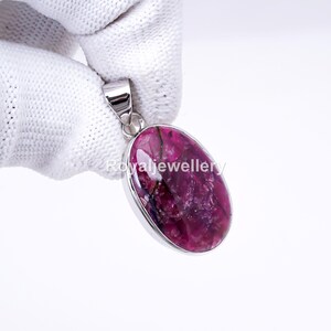 Op de afbeelding: Een zilveren hanger met een ovale, dieproze edelsteen. De edelsteen heeft een textuur met donkere aderen. De hanger wordt vastgehouden door een gehandschoende hand tegen een witte achtergrond. De tekst "Royaljewellery" is zichtbaar.
