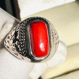 May include: A silver ring featuring a vibrant, rectangular red stone. The ring's band and setting are intricately patterned. The ring is displayed against a white background.