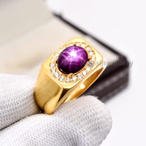 Può includere: Un anello d'oro con una grande gemma ovale, viola scuro, con effetto stella, circondata da piccole gemme trasparenti. L'anello ha una faccia rettangolare e una fascia in oro lucido. L'anello è esposto su una superficie bianca.