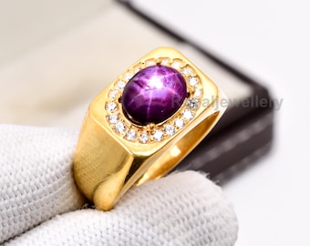 May include: A gold ring with a large, oval, deep purple gemstone with a star effect, encircled by small, clear gemstones. The ring has a rectangular face and a polished gold band. The ring is displayed on a white surface.