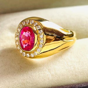 Puede incluir: Un anillo de oro con una gran gema rosa brillante de corte ovalado, rodeada por un halo de pequeñas gemas claras. La banda del anillo es gruesa y pulida. El anillo está sobre un fondo neutro.