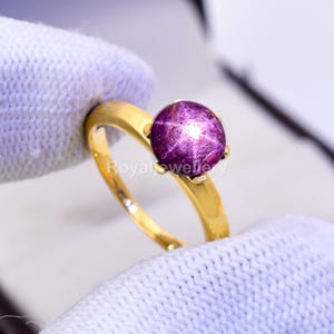 May include: A gold ring with a large, round star ruby gemstone. The ruby is a deep purple colour with a star effect. The ring is held by a gloved hand, showing the polished gold band.