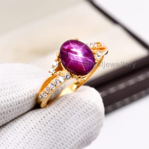 May include: A gold ring featuring a large, oval, deep-red gemstone with a star-like effect. The ring is adorned with small, clear gemstones along the band and around the main stone, creating a luxurious look.