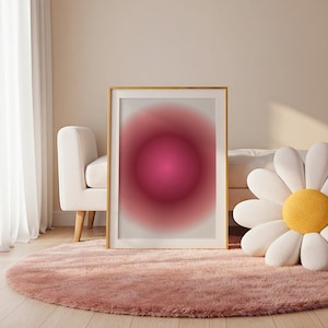 Puede incluir: Una impresión artística enmarcada con un diseño abstracto. La obra presenta un degradado circular rosa oscuro central que se desvanece a un rosa más claro. La impresión se muestra en una pared de color claro, sobre una alfombra rosa y esponjosa.