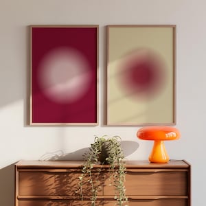 Könnte beinhalten: Zwei gerahmte Kunstdrucke mit abstrakten, verschwommenen Kreisen in Rot- und Cremetönen. Eine orangefarbene pilzförmige Lampe steht auf einer Holzkommode unter den Drucken. Eine Topfpflanze mit Ranken befindet sich ebenfalls auf der Kommode.