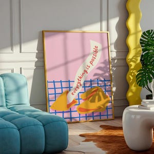 Op de afbeelding: Een ingelijste kunstafdruk met een roze achtergrond, een blauw rasterpatroon en een illustratie van een fortune cookie. De tekst op het papiertje luidt "everything is possible". De afdruk zit in een gouden lijst.