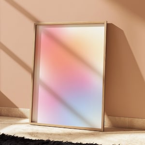 Puede incluir: Una impresión artística enmarcada con un suave degradado de colores rosa, naranja, amarillo y azul. La obra de arte está en un marco de madera clara y se apoya en una pared color melocotón, con sombras proyectadas.