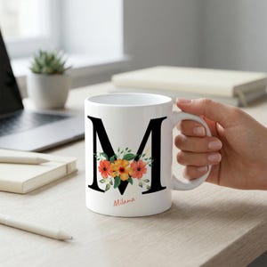 May include: A white ceramic mug featuring a large black capital letter M decorated with a cluster of orange and yellow watercolor flowers and green leaves. The name Milana is printed in a small red script font directly below the letter M.